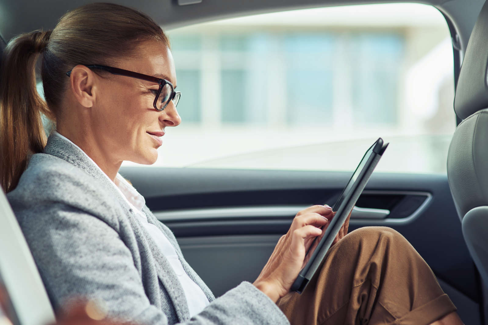 Side of a successful smiling business woman wearing eyeglasses using digital tablet, working while sitting on a taxi back seat