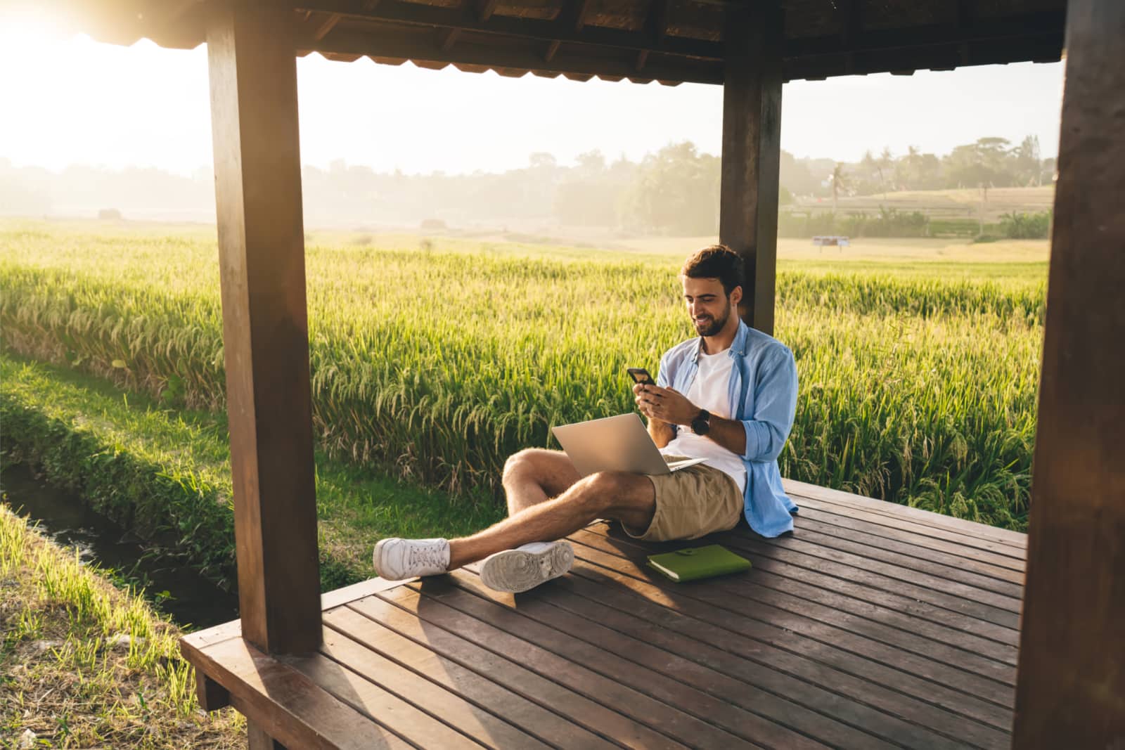 Jovem empresário sentado numa varanda com vista para um campo de milho enquanto usa o laptop e o smartphone